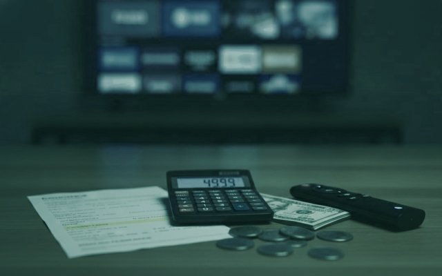 A calculator, dollar bills, coins, a streaming stick and a remote on a table in front of a TV